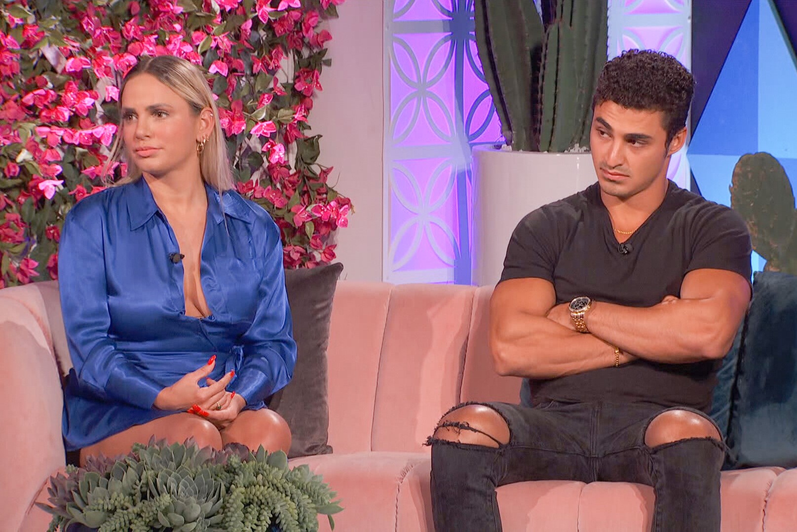 Indy Santos and Joseph Abdin, who starred in 'Big Brother 24' on CBS, sit next to each other on a couch during the jury round table. Indy wears a royal blue long-sleeved dress. Joseph wears a black shirt and black jeans with ripped knee holes.
