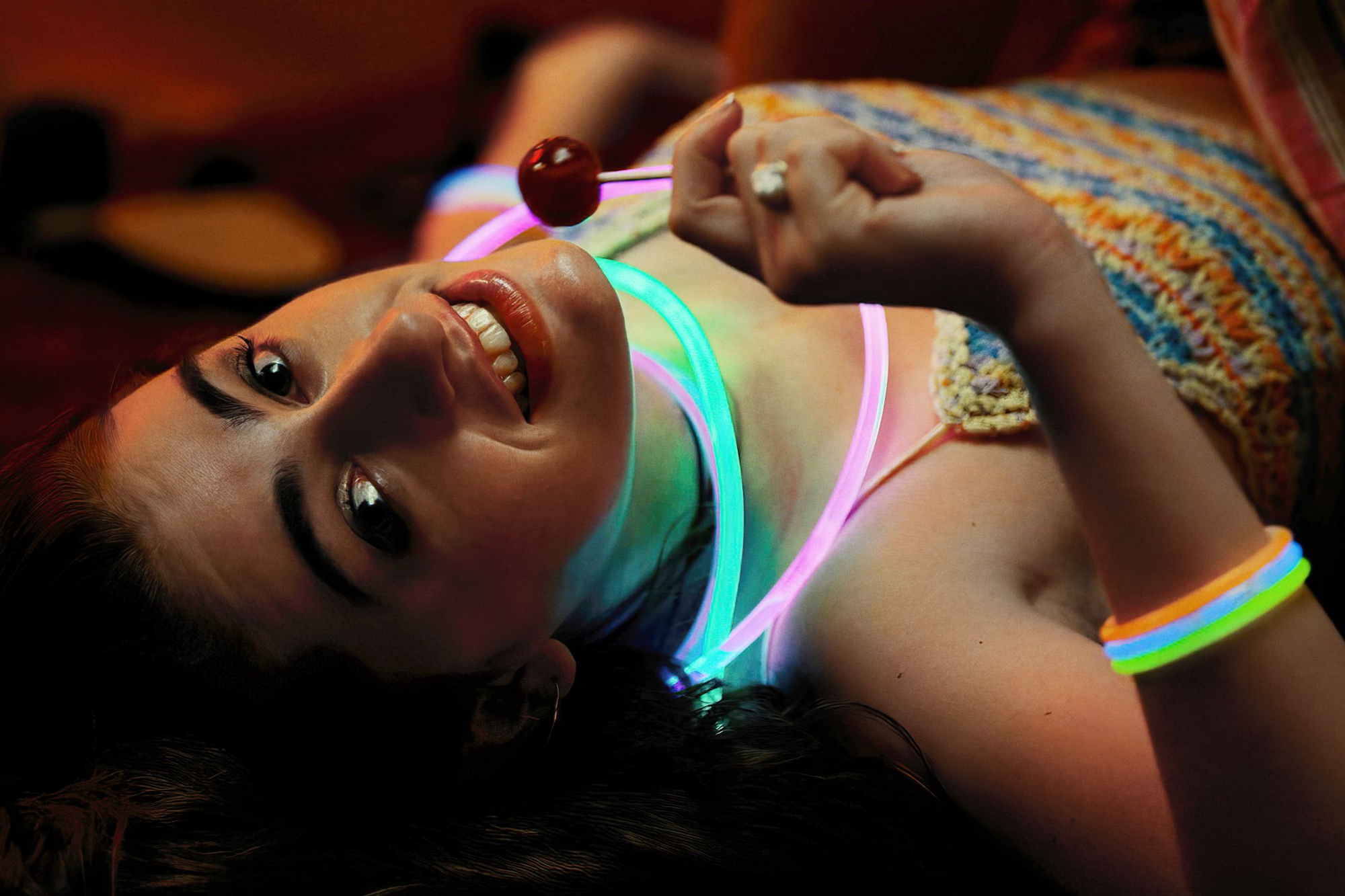 'Bodies Bodies Bodies' Rachel Sennott as Alice laying on her back, looking up at the camera wearing neon glow sticks around her neck and holding a lollipop