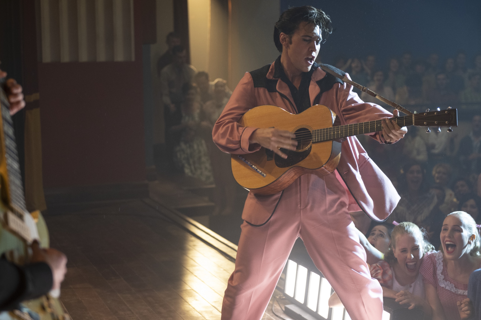 'Elvis' Austin Butler as Elvis Presley wearing a pink and black outfit, playing the guitar on stage in front of a bunch of fans.