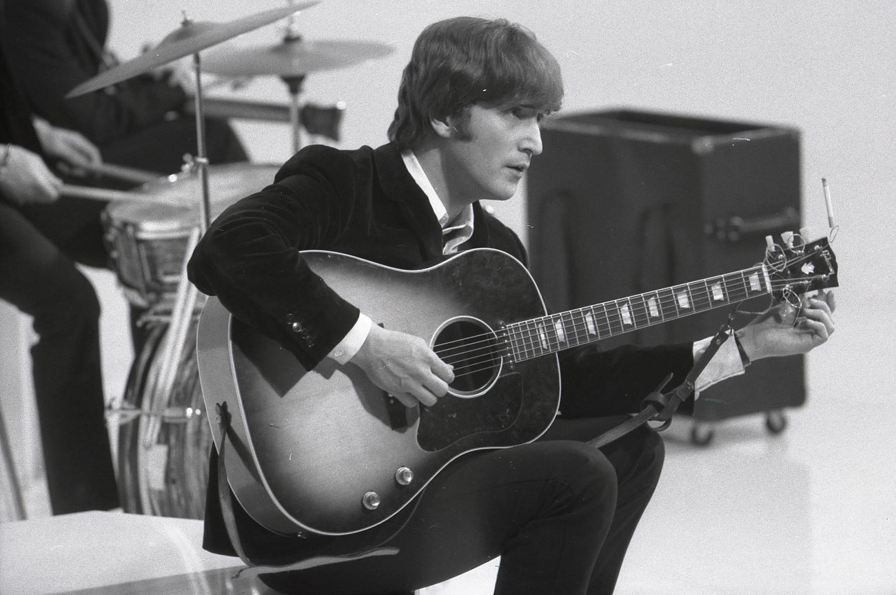 A black and white picture of John Lennon sitting and holding a guitar.