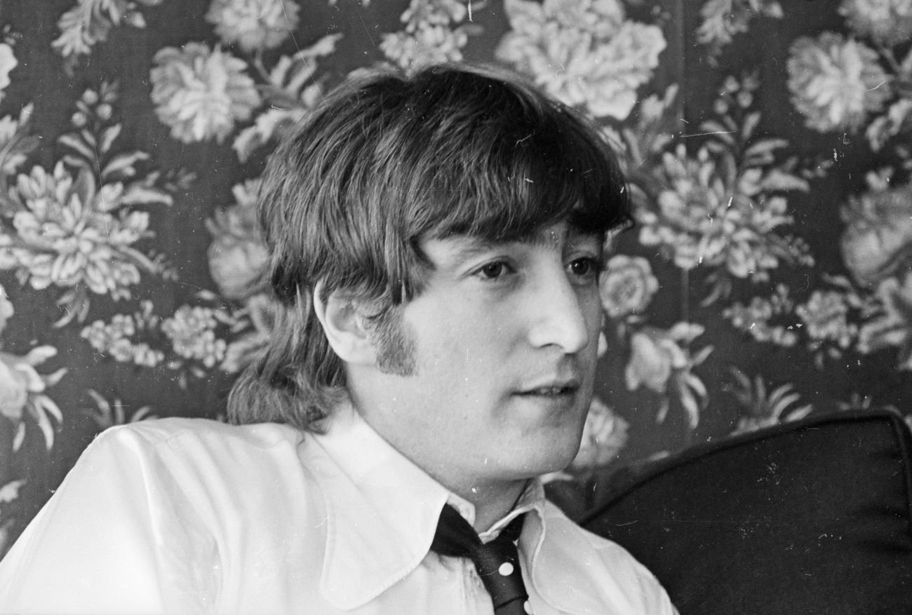 A black and white picture of John Lennon sitting in front of a floral wallpaper.