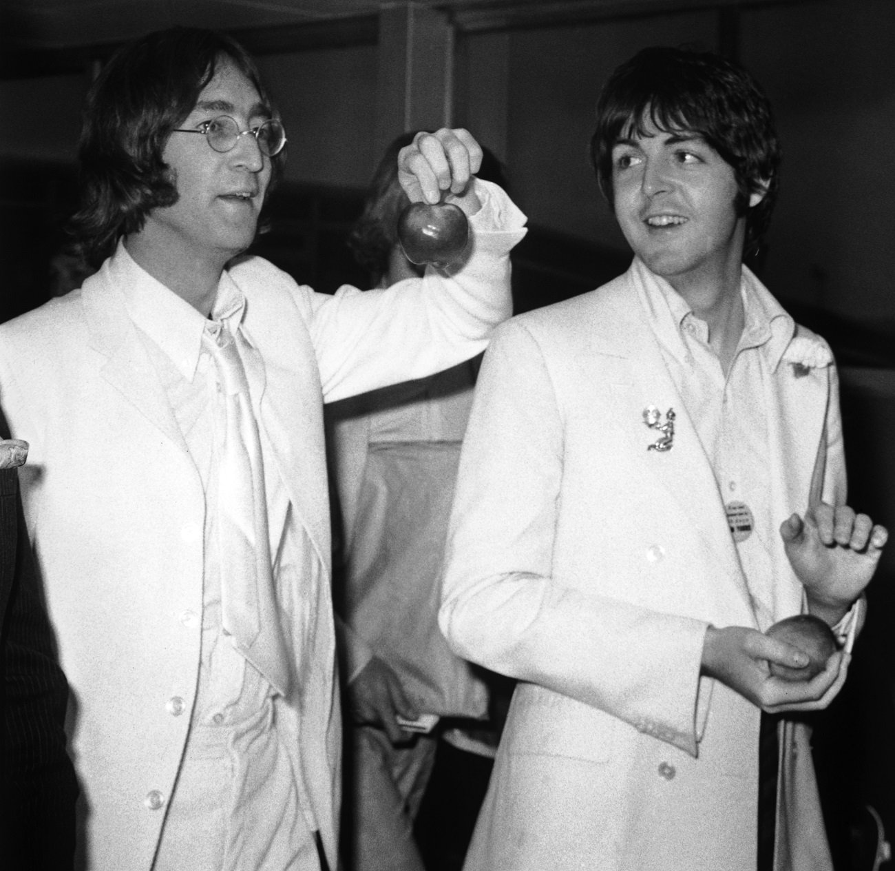 A black and white picture of John Lennon and Paul McCartney wearing white suits and holding apples. 