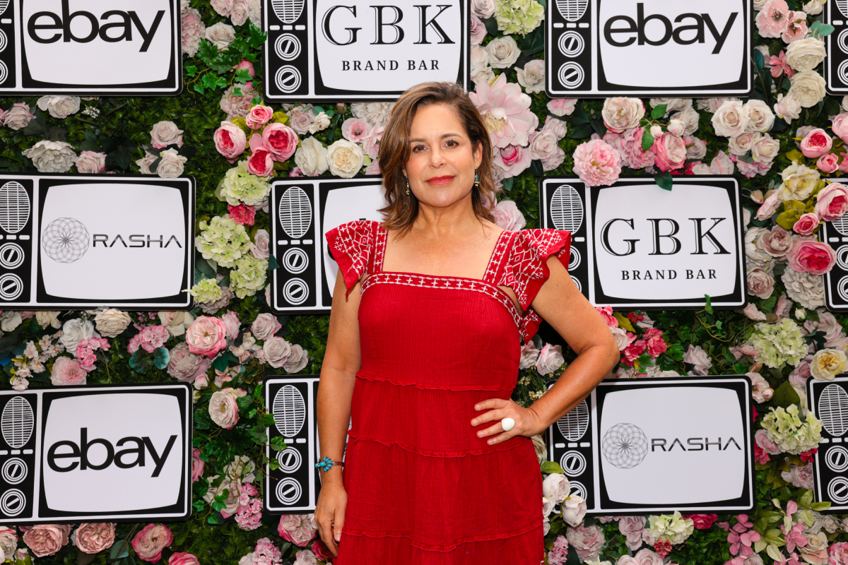 The Patient actor Laura Niemi wears a red dress and poses in front of a wall of flowers. 