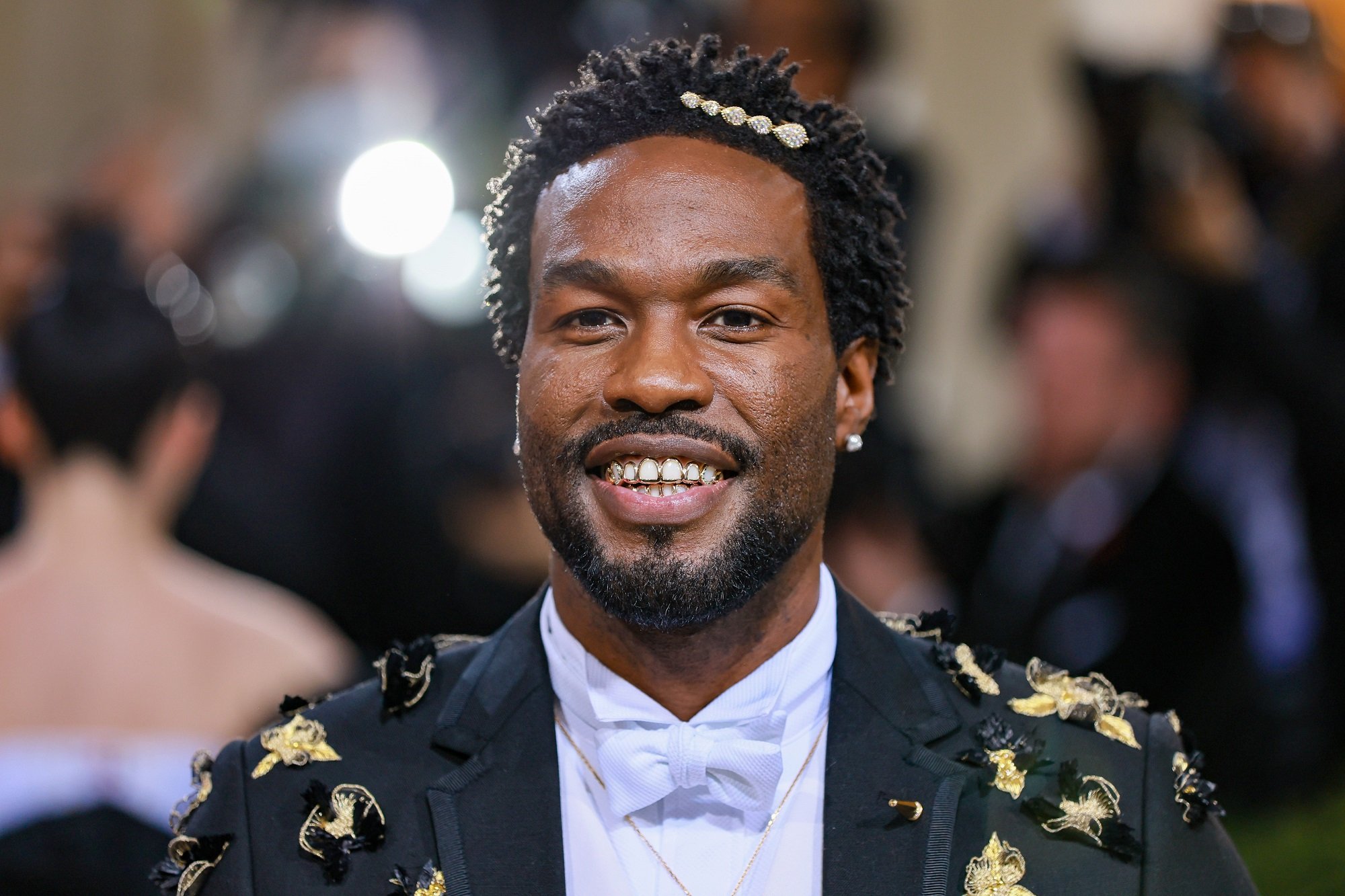 Yahya Abdul-Mateen II smiles at the camera while wearing a black suit with gold and black ribbons