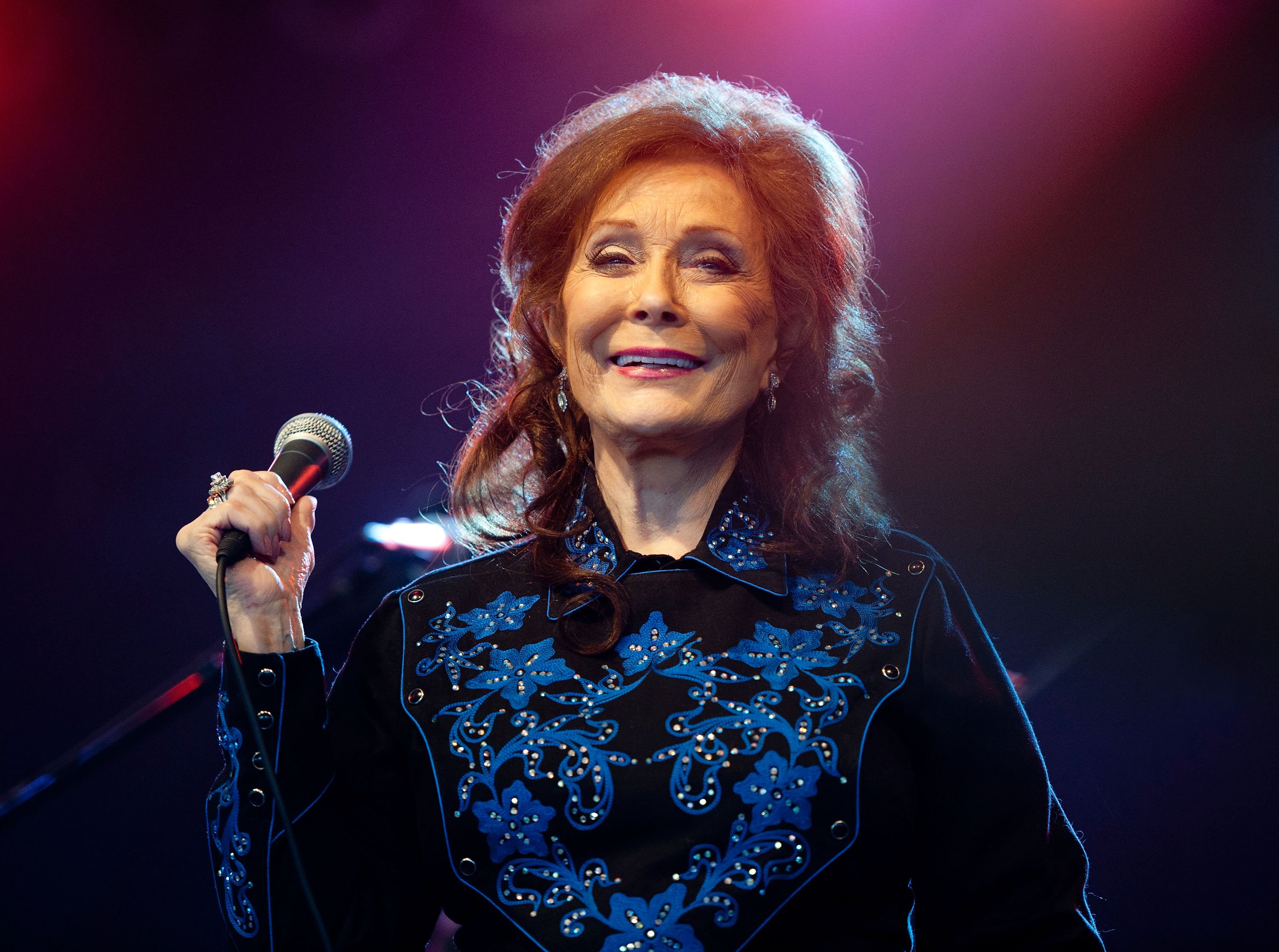 Loretta Lynn holds a microphone while wearing a black-and-blue shirt