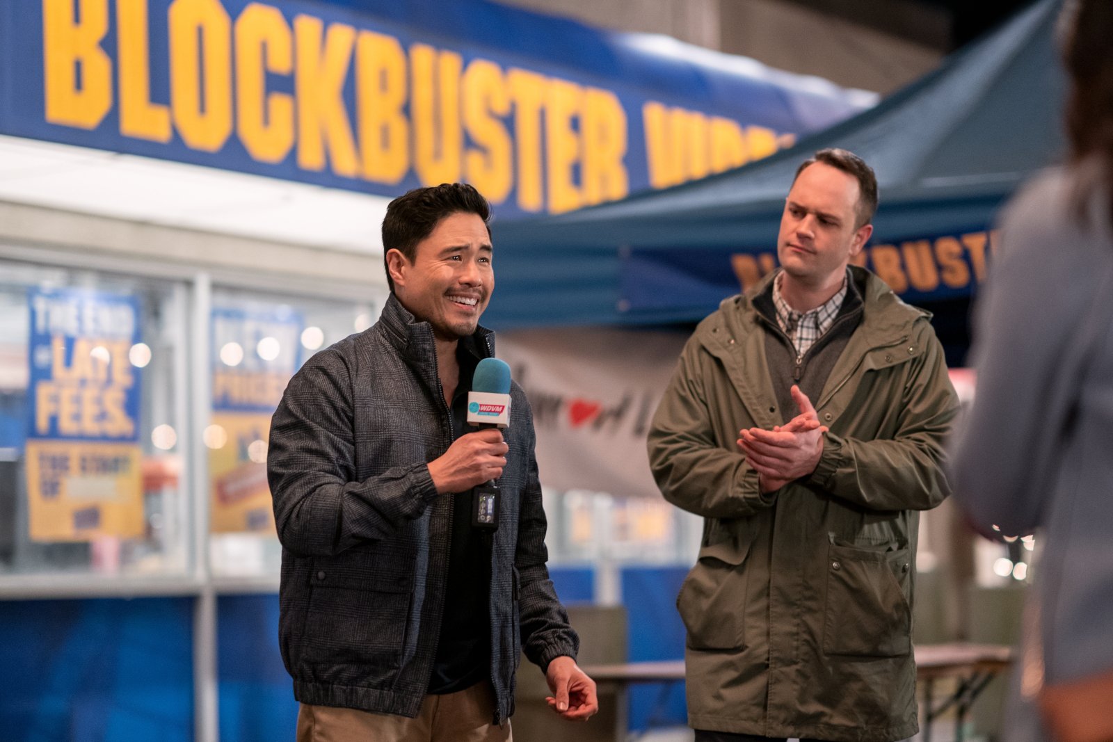 Randall Park and Simon Druker as Timmy Yoon and Remington Alexander in 'Blockbuster' on Netflix for our article about what's true. Timmy is holding a microphone and they're standing in front of the store.