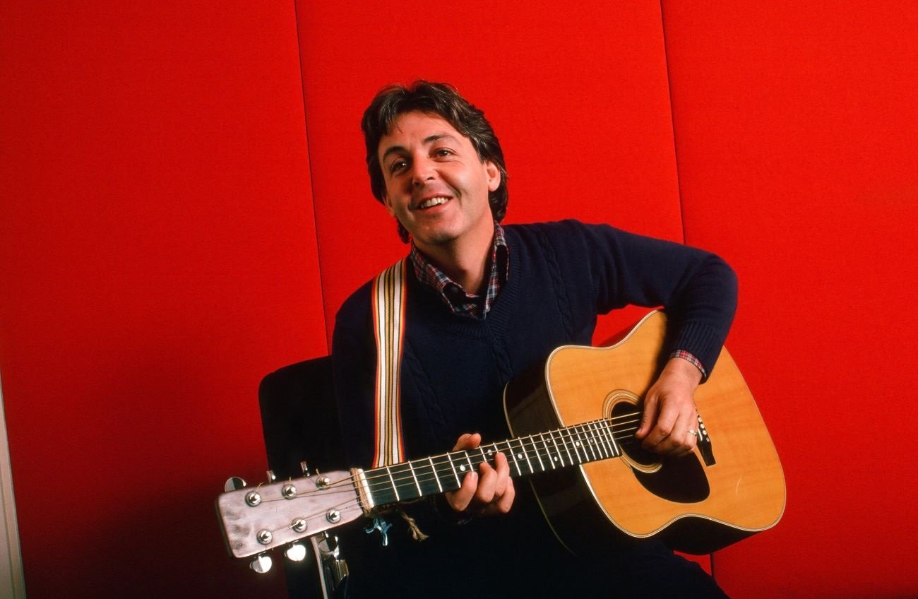 Paul McCartney sits and holds a guitar against a red background. 