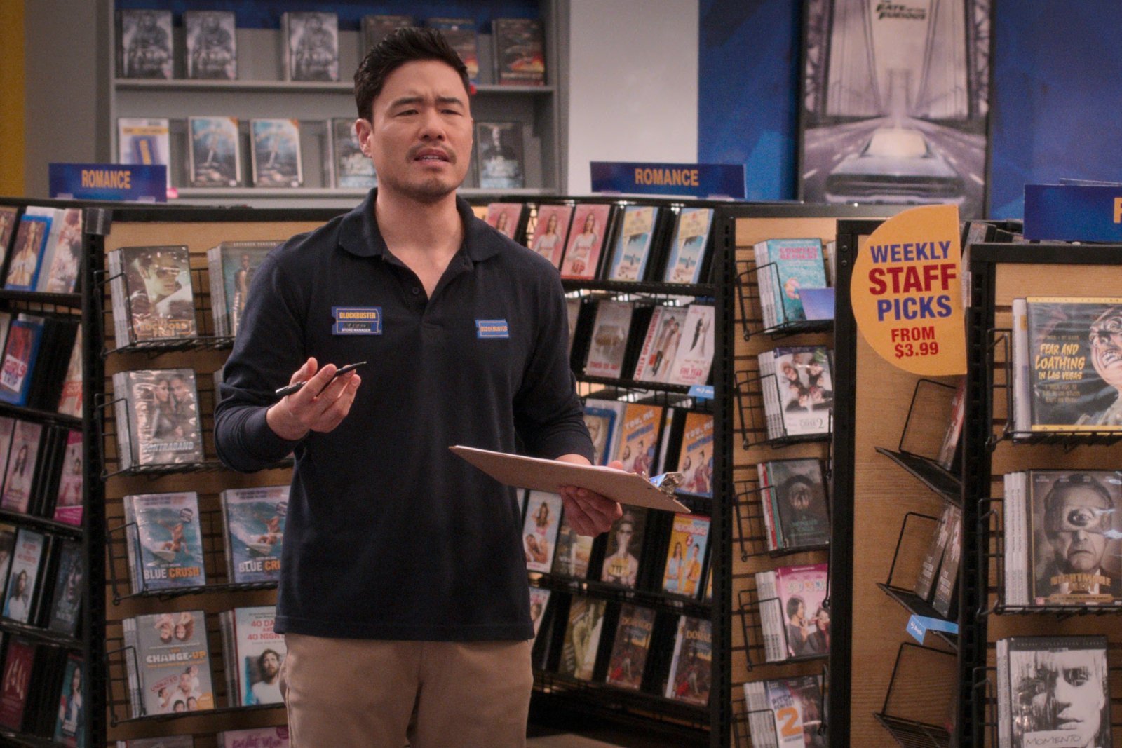 Randall Park as Timmy in 'Blockbuster' on Netflix. He's wearing a Blockbuster uniform, standing in front of video displays, and he's holding a clipboard.