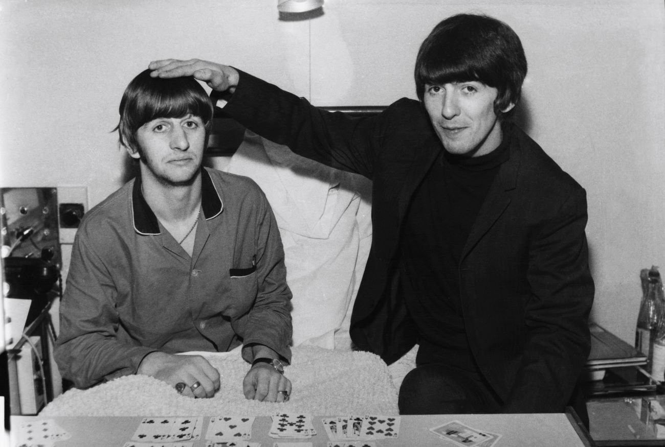 A black and white picture of Ringo Starr sitting in bed and George Harrison patting him on the head.