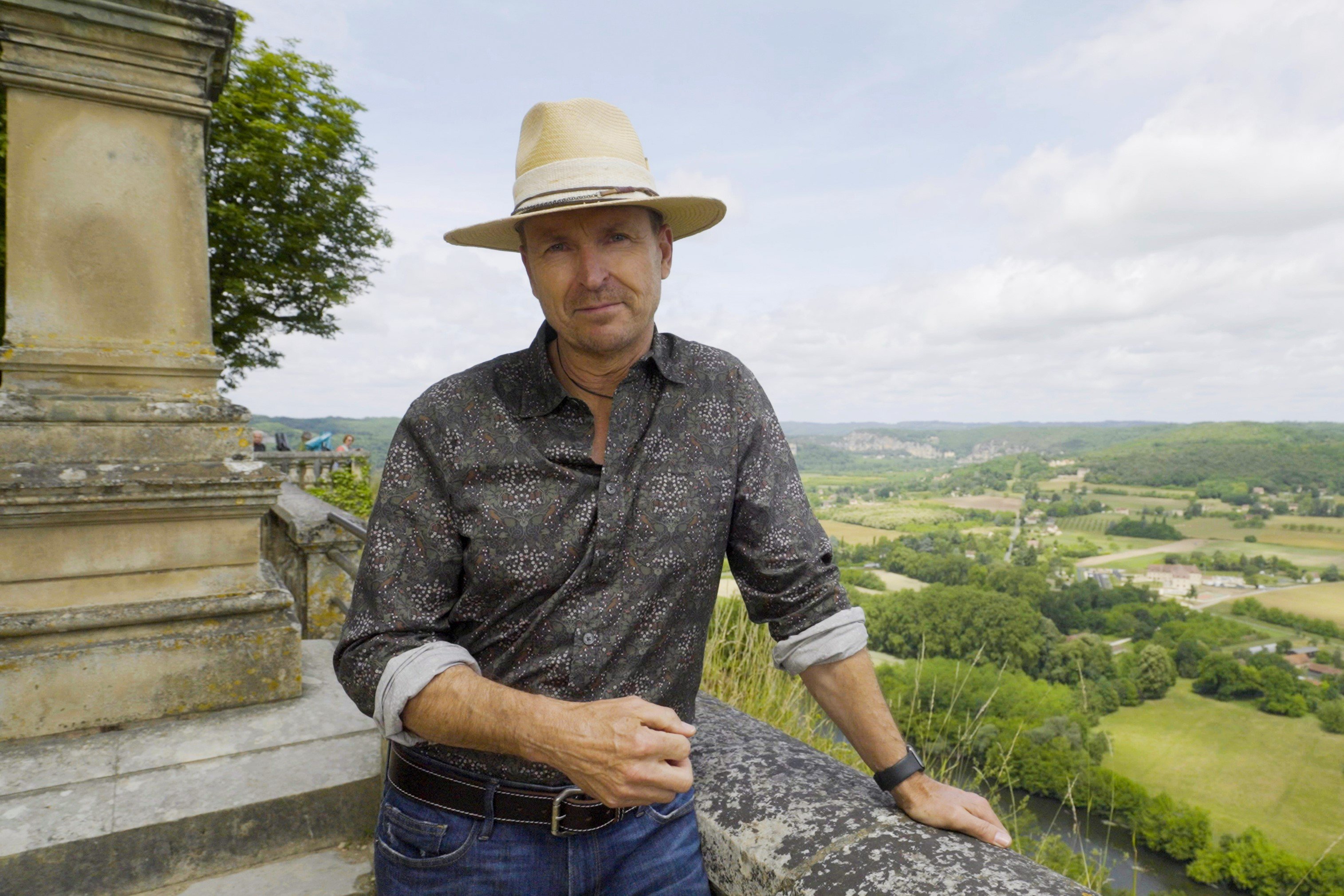 Phil Keoghan, who hosts 'The Amazing Race' Season 34 on CBS, wears a dark gray patterned button-up long-sleeved shirt, jeans, and a tan hat.