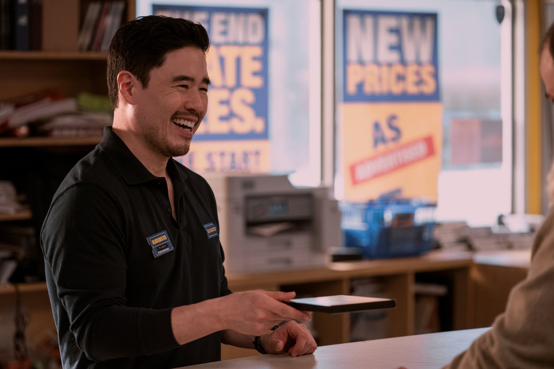 Randall Park in Netflix's new show, 'Blockbuster,' for our article about what time it premieres. He's wearing a Blockbuster uniform, standing in front of a counter, and holding out a DVD.