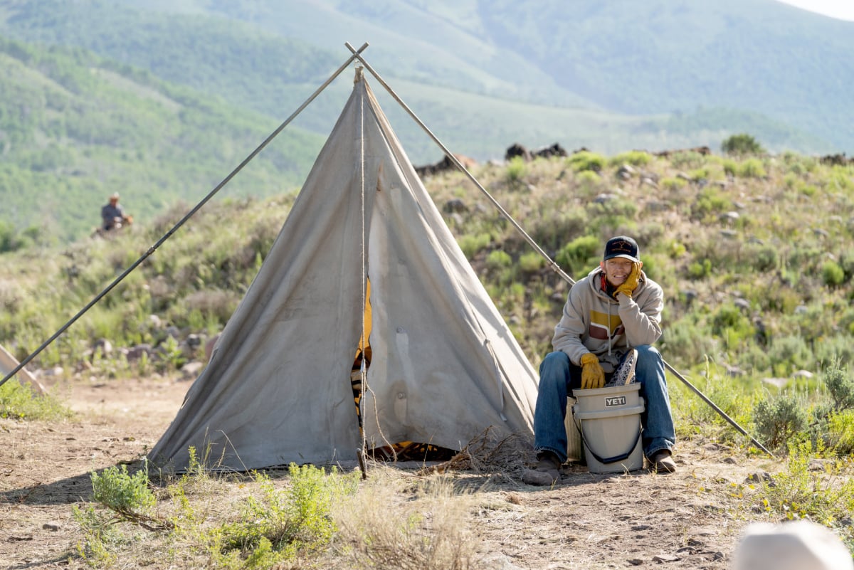 In Yellowstone, Jimmy sits on a bucket outside of a tent.