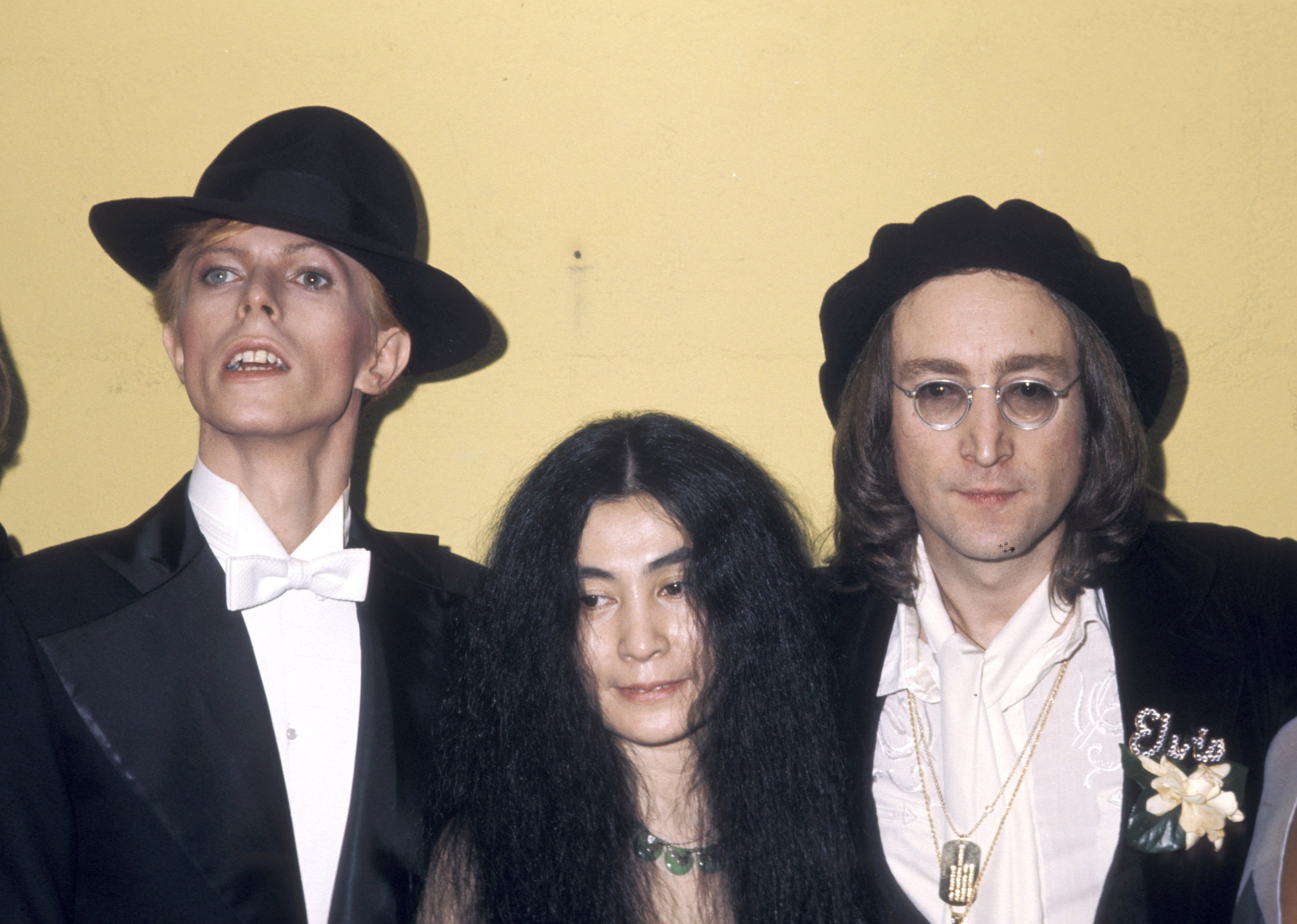 David Bowie, Yoko Ono, and John Lennon wear black and pose against a yellow background. 