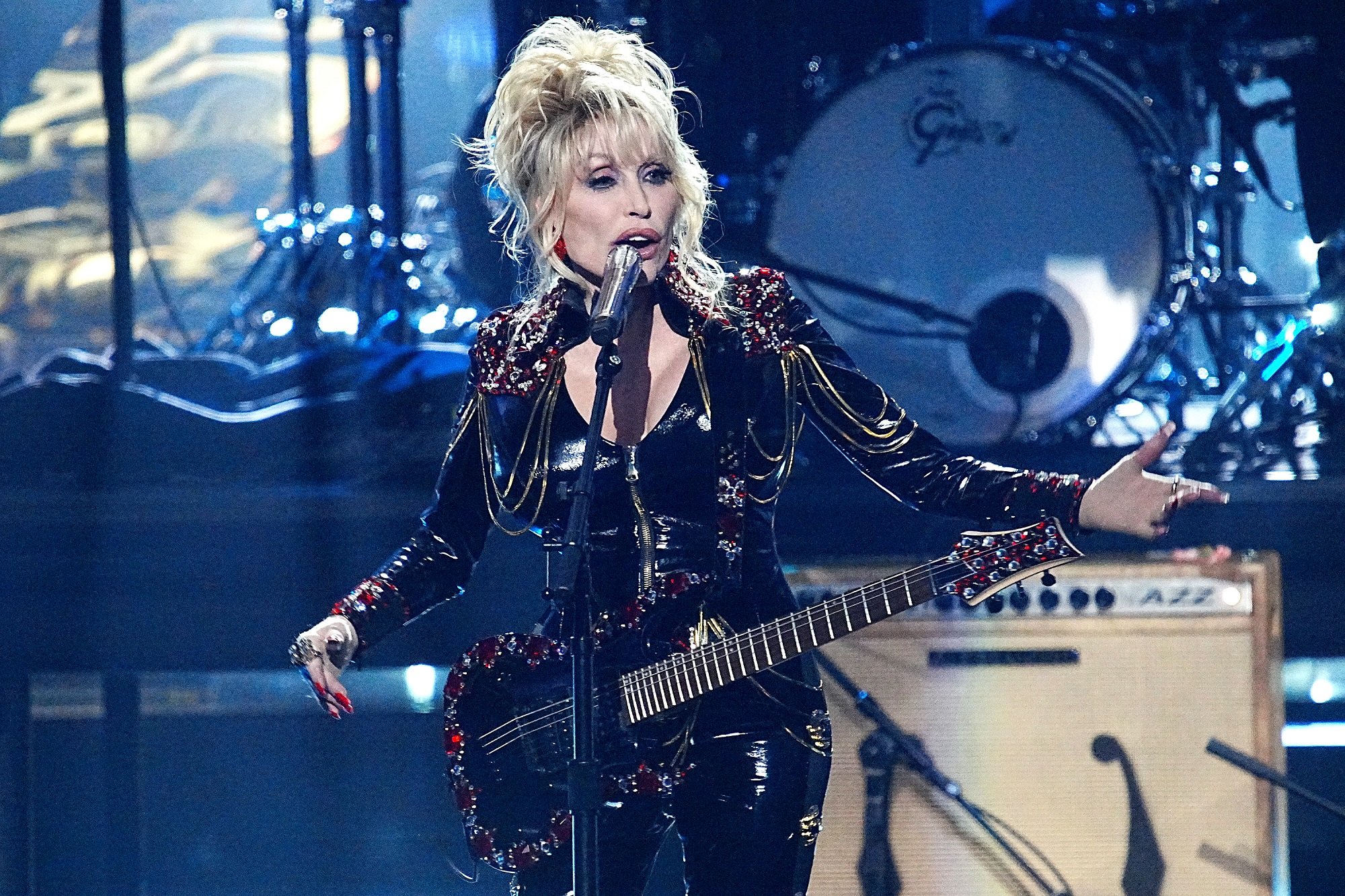 Dolly Parton performs on stage with a black guitar while wearing a black outfit