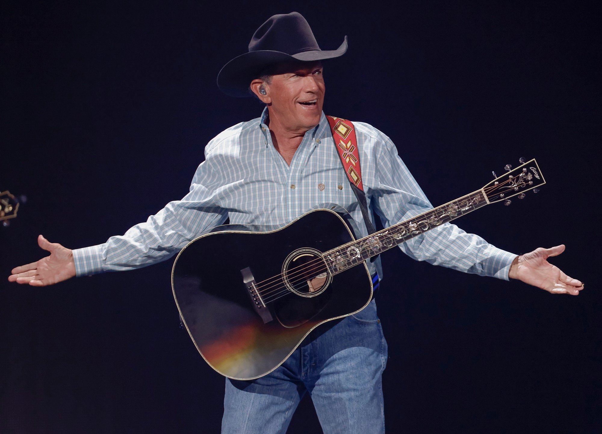 George Strait smiles with a guitar around his neck and holds his arms stretched out on stage