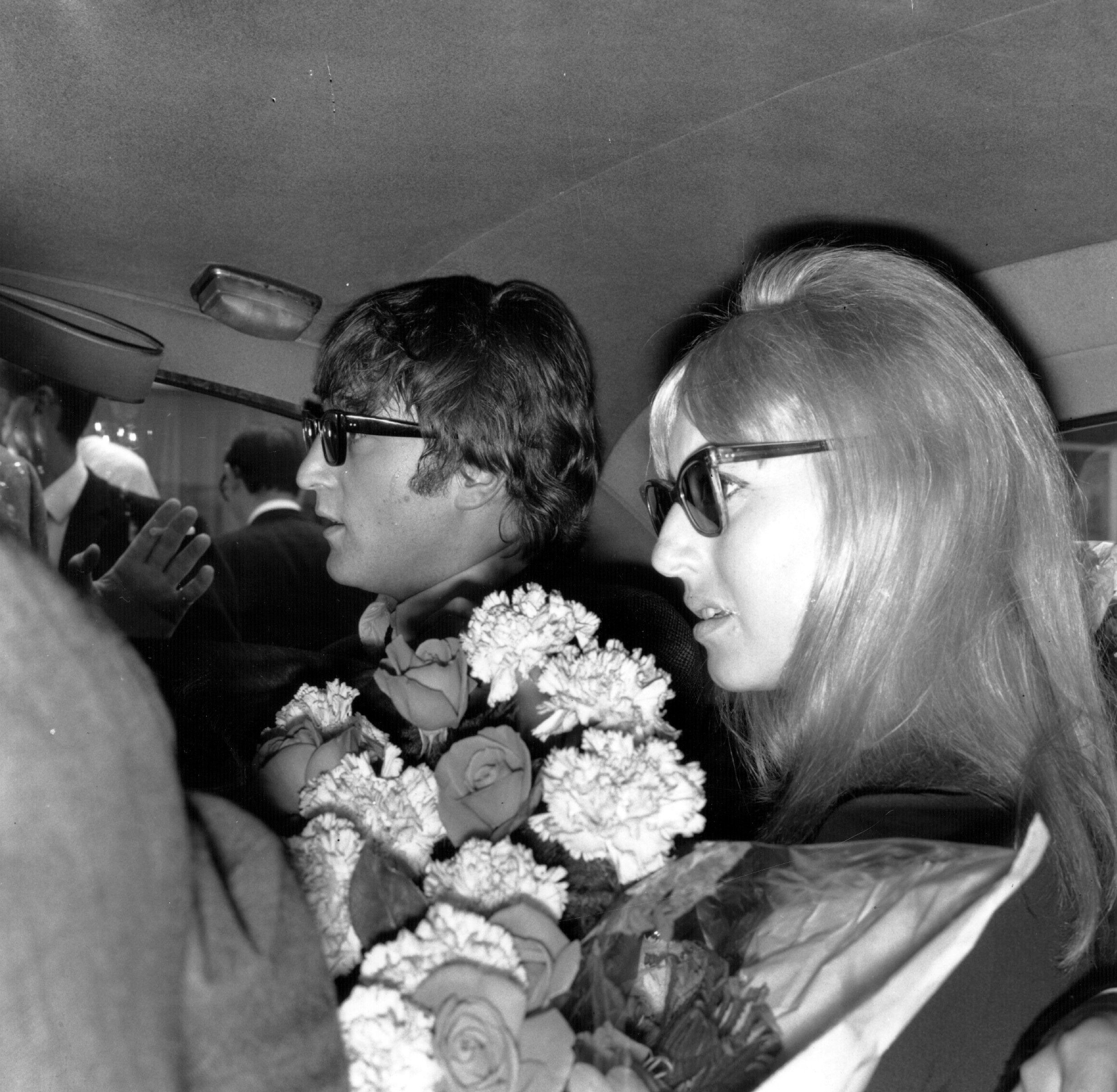 A black and white picture of John and Cynthia Lennon sitting in the backseat of a car holding flowers.