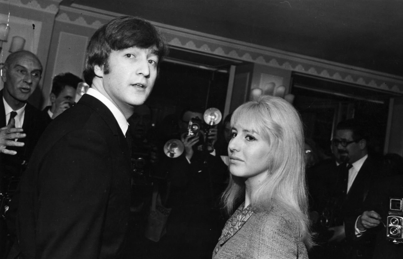 A black and white picture of John Lennon and his wife Cynthia Lennon standing in front of photographers.