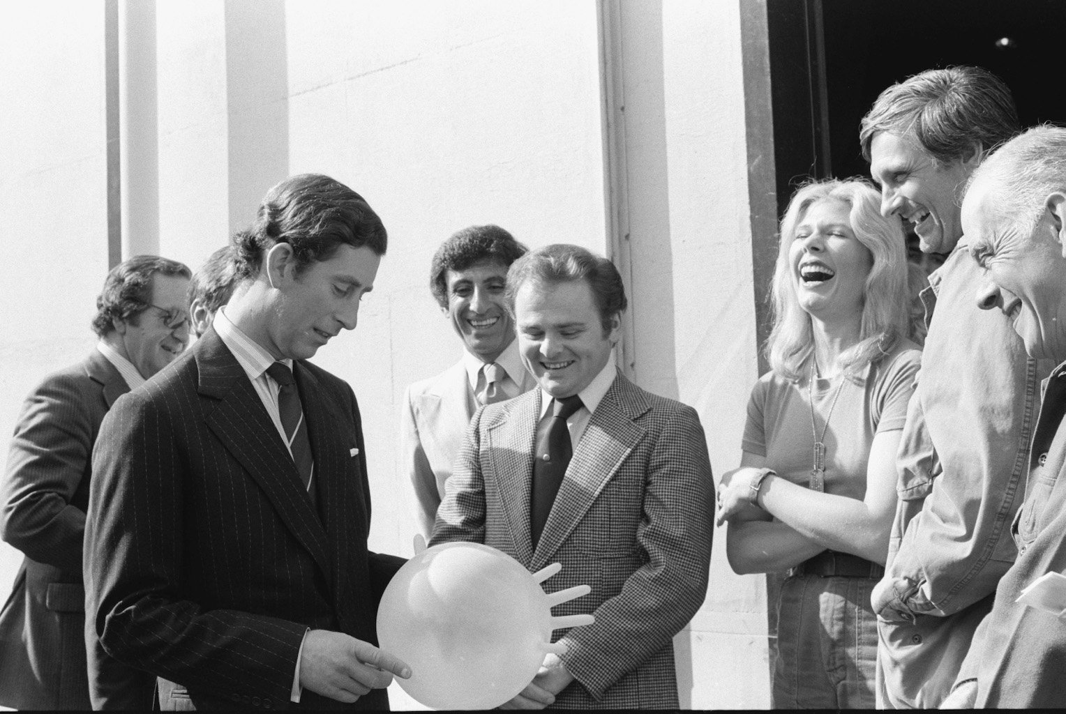 A black and white photo of Prince Charles on the set of 'M*A*S*H' laughing with the cast while holding an inflated surgical glove