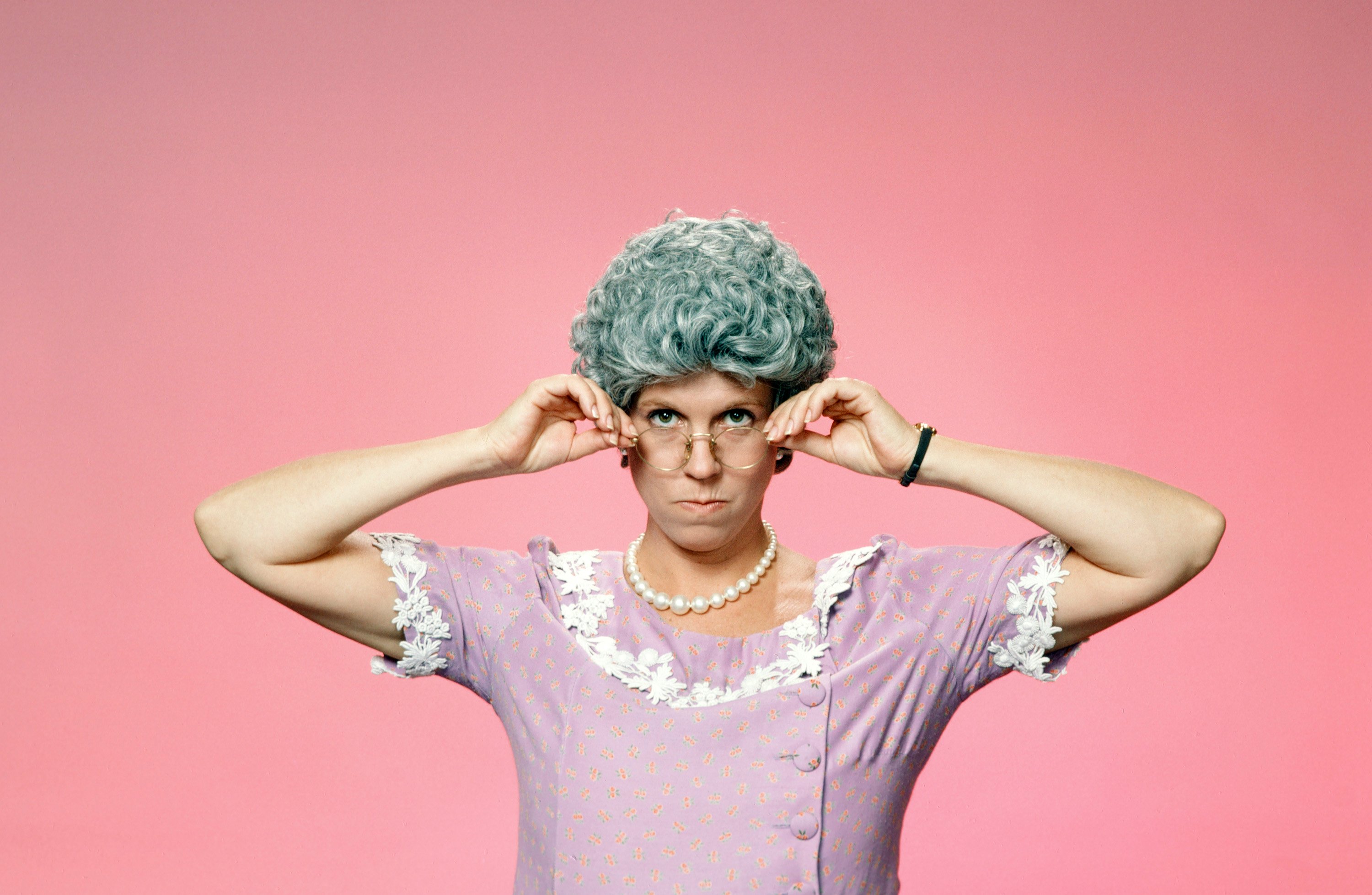 'Mama's Family' star Vicki Lawrence in a purple dress, poses as her character Thelma Harper in front of pink backdrop.