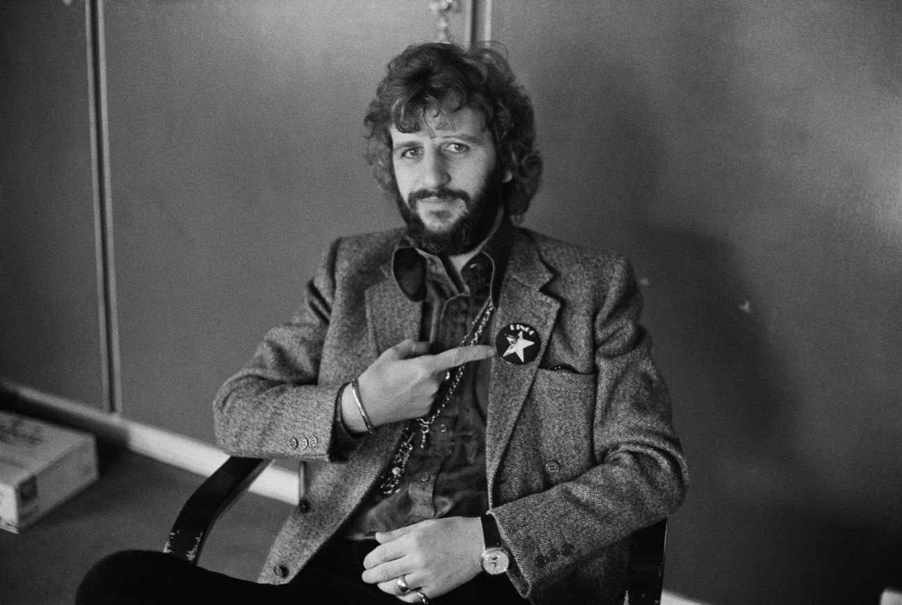 A black and white picture of Ringo Starr sitting in a chair and pointing to a star sticker on his jacket.