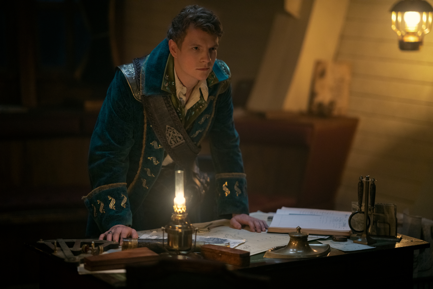 Patrick Gibson as Sturhmond in 'Shadow and Bone' Season 2. He's leaning over a table with books and papers on it.