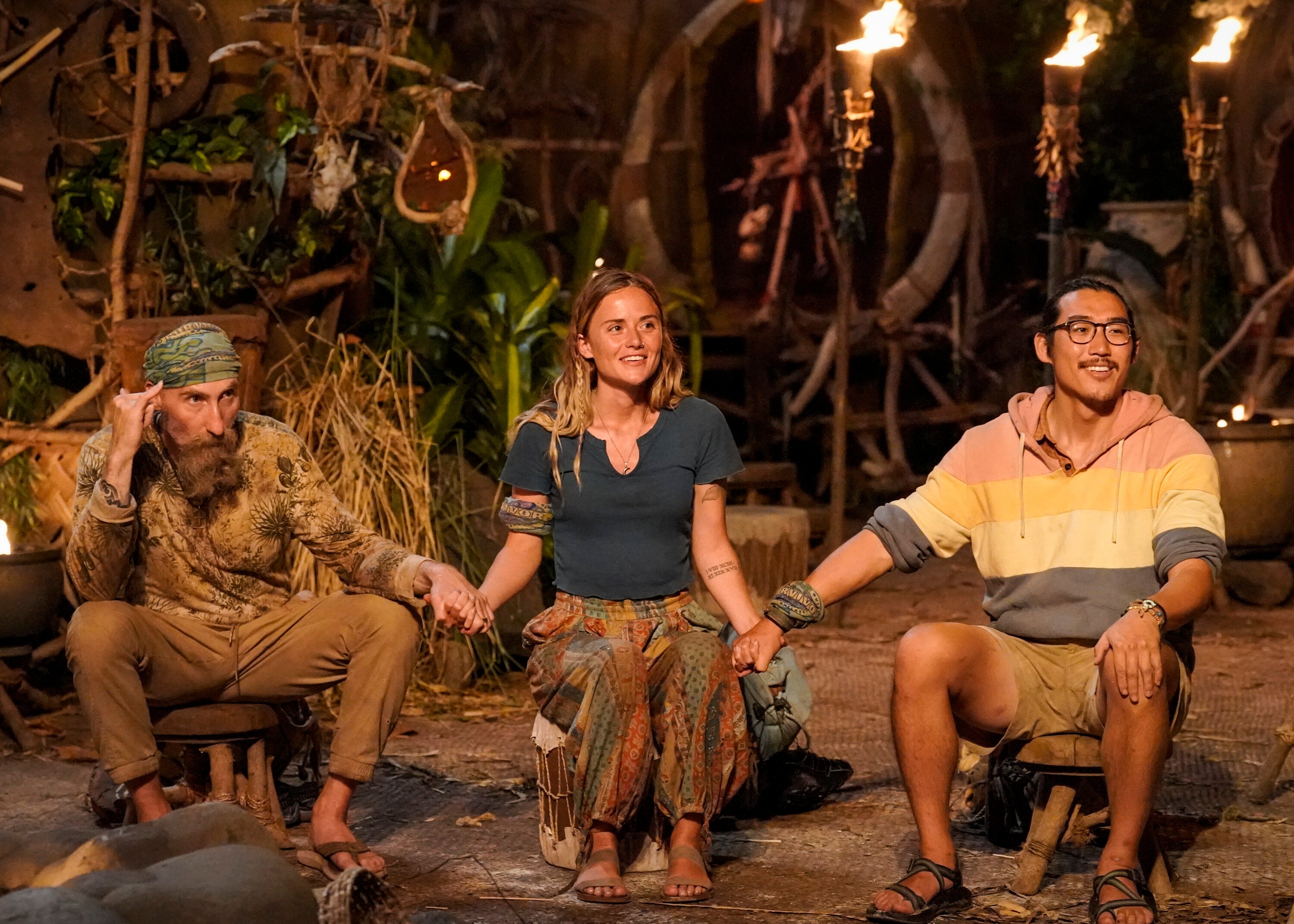 Mike Gabler, Cassidy Clark, and Owen Knight hold hands during the Final Tribal Council in 'Survivor' Season 43 on CBS. Gabler wears a camo shirt and tan pants. Cassidy wears a dark blue shirt and multi-colored pants. Owen wears a pink, yellow, white, and blue striped hoodie and tan shorts.