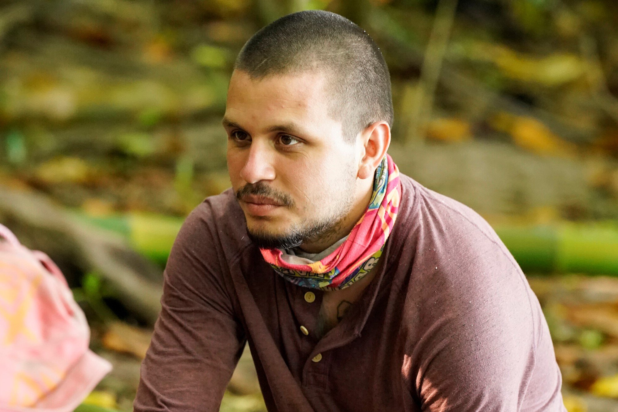 Jesse Lopez, who stars in 'Survivor' Season 43, wears a faded dark red henley shirt and his red 'Survivor' buff around his neck.