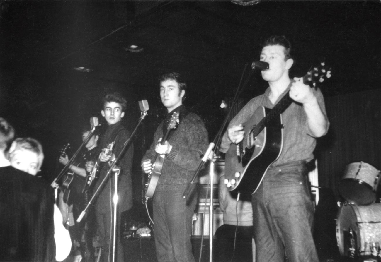 A black and white picture of Beatles George Harrison and John Lennon onstage in Germany with Tony Sheridan.