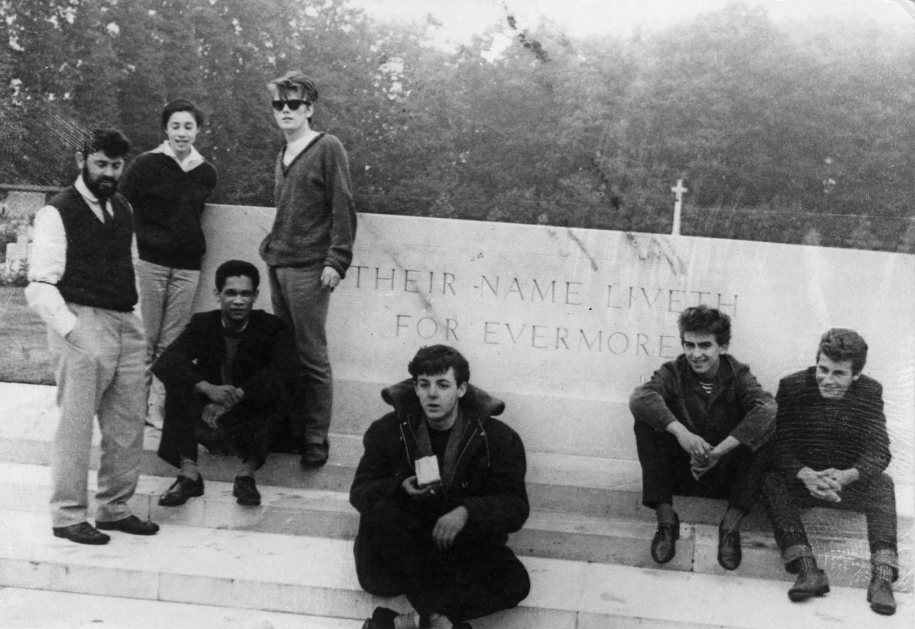 A black and white picture of the early Beatles, Stuart Sutcliffe, Paul McCartney, George Harrison, and Pete Best, and Allan Williams, Beryl Williams, and musician Lord Woodbine, posing on steps.  