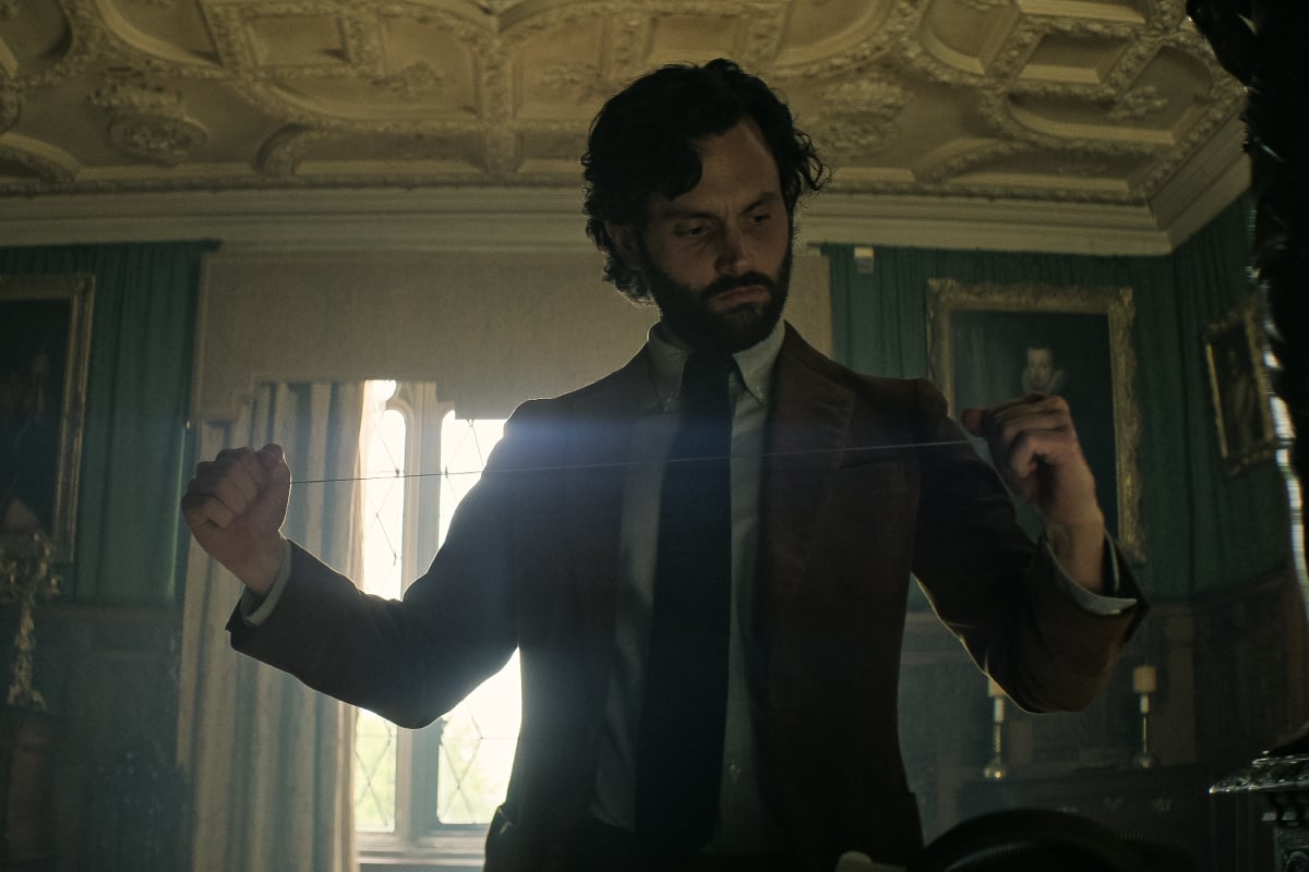 Penn Badgley as Joe Goldberg in 'You' Season 4. Joe wears a suit and tie, has a beard and longer hair, and stretches out a piece of string. 