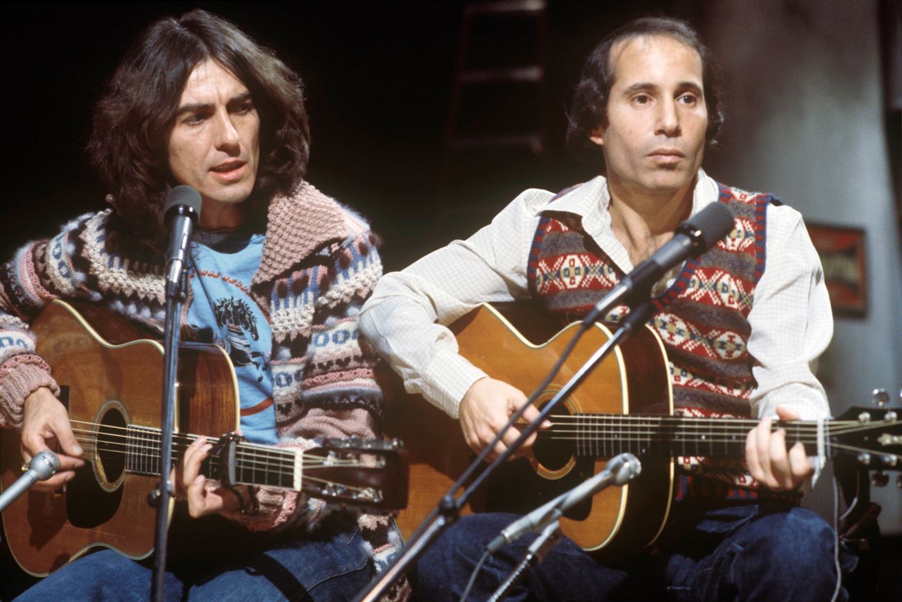 George Harrison and Paul Simon sit next to each other holding guitars.