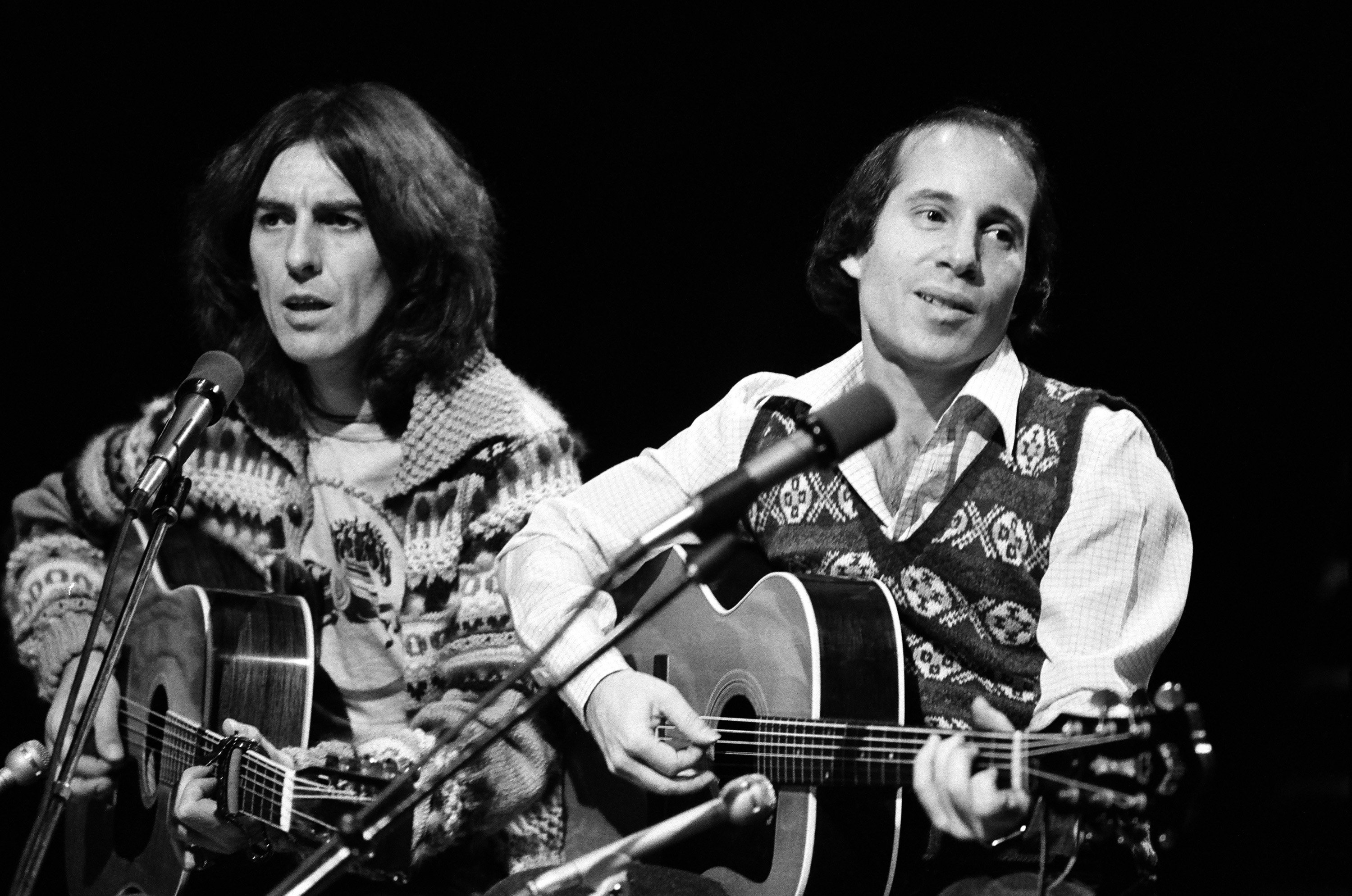 A black and white picture of George Harrison and Paul Simon holding guitars and sitting in front of microphones.