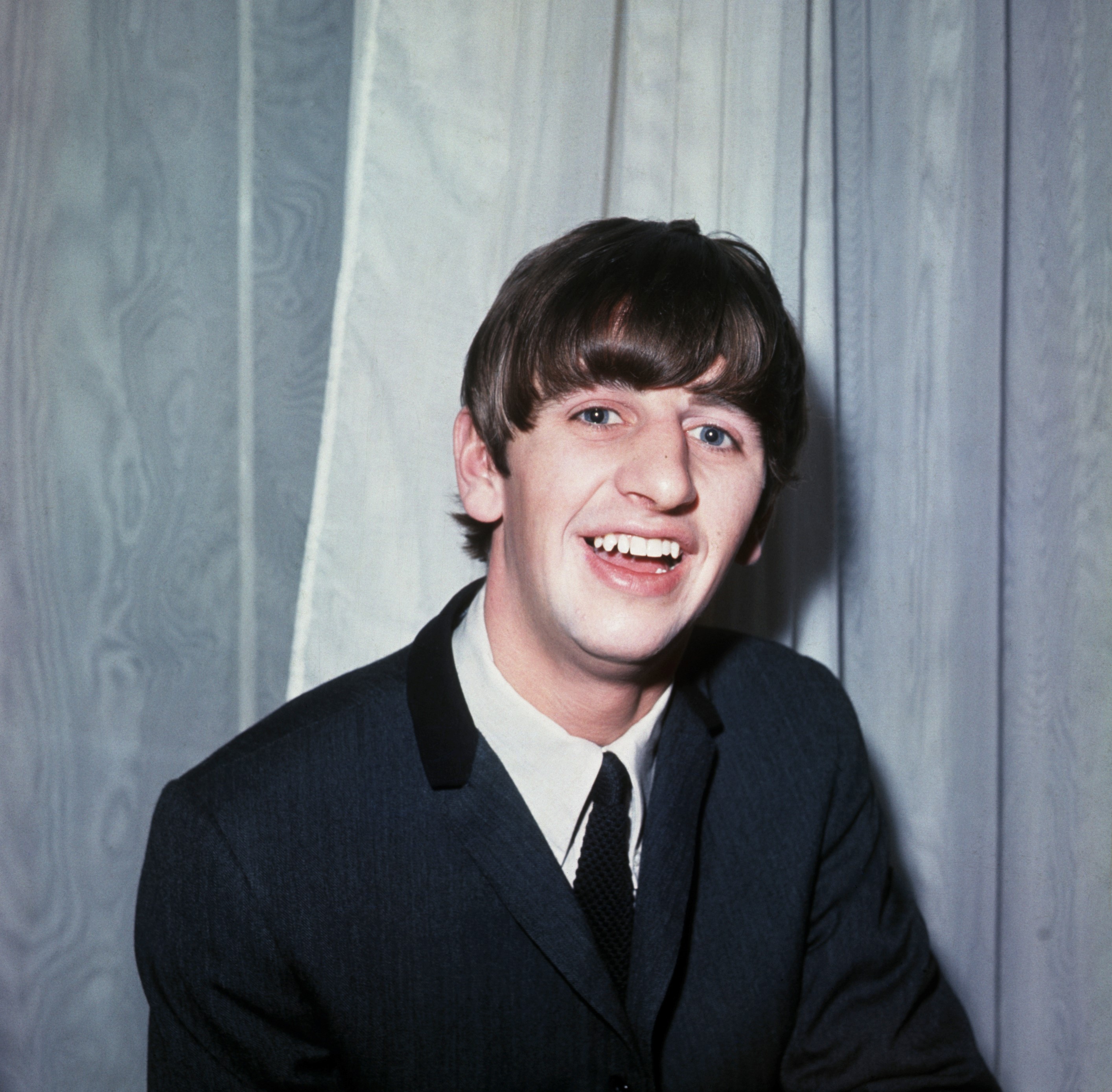 Ringo Starr wears a suit and sits against a wood paneled background.