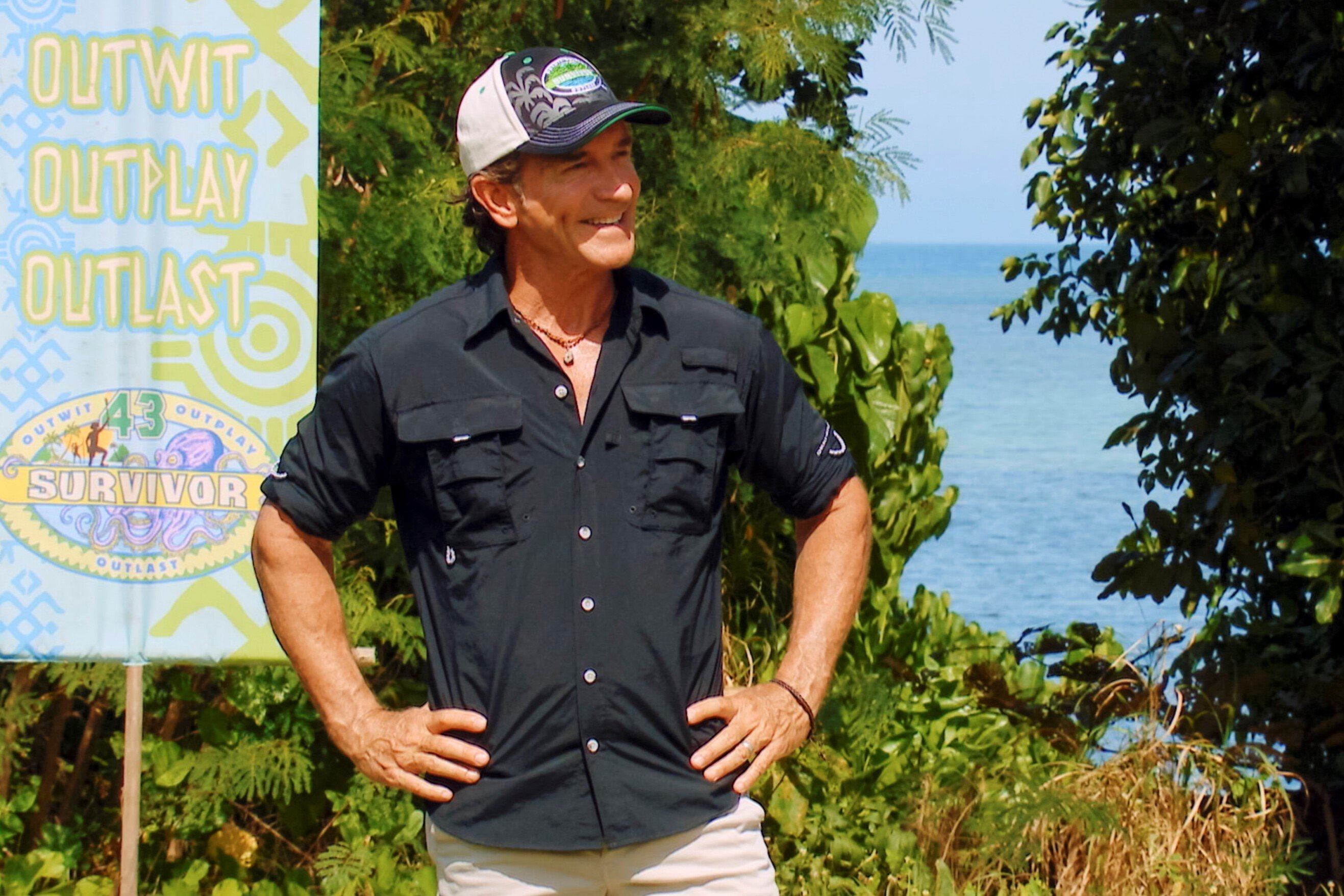 Jeff Probst, who hosts the reality competition series, 'Survivor,' on CBS, wears a black button-up shirt with rolled-up sleeves, white shorts, and a black, white, blue, and green 'Survivor' baseball hat.