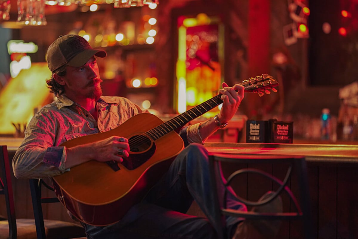 The Tulsa King Season 1 Finale features Garrett Hedlund singing. Mitch sits on a barstool holding his guitar.