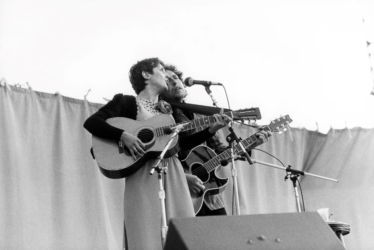 A black and white picture of Joan Baez and Bob Dylan playing guitars together in front of microphones.