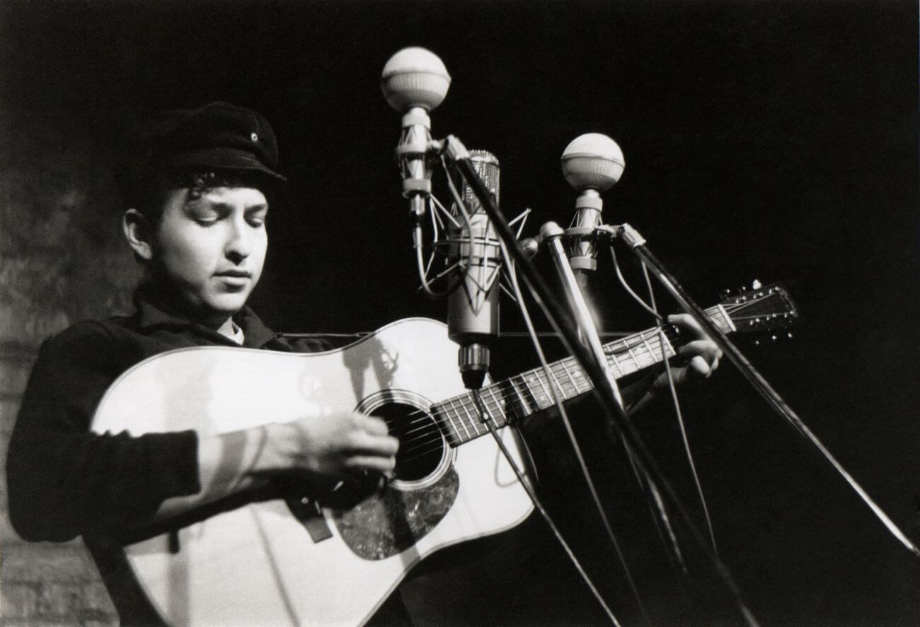 A black and white picture of Bob Dylan playing guitar in front of multiple microphones.