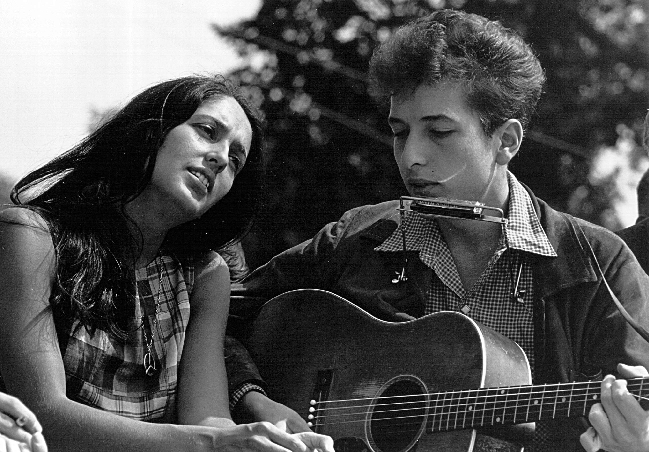 A black and white picture of Joan Baez sitting next to Bob Dylan, who plays guitar.