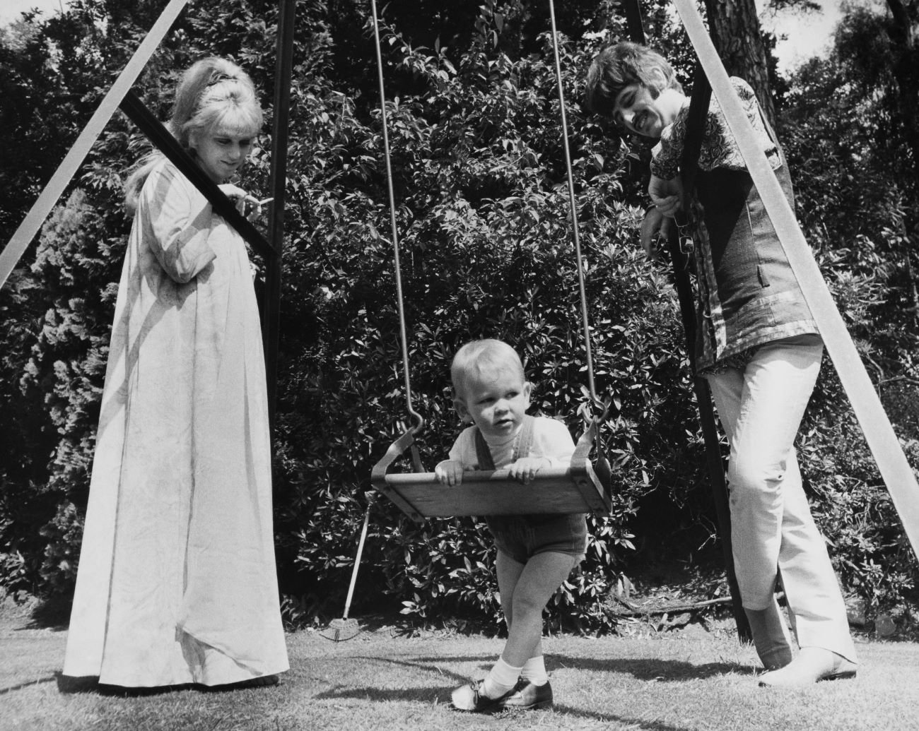 A black and white picture of Maureen Starkey and Ringo Starr looking down at their son Zak on a swing.