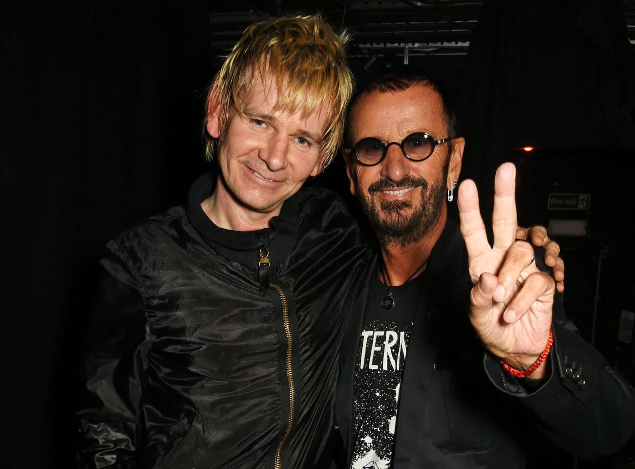 Ringo Starr's son Zak Starkey stands with his arm around his father's shoulders. Ringo Starr holds up a peace sign. 