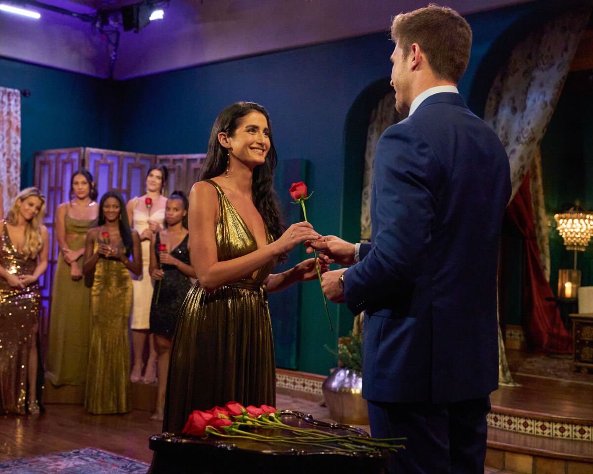 During a rose ceremony, The Bachelor Zach Shallcross hands Ariel Frenkel a rose. She wears a gold dress and earrings. 