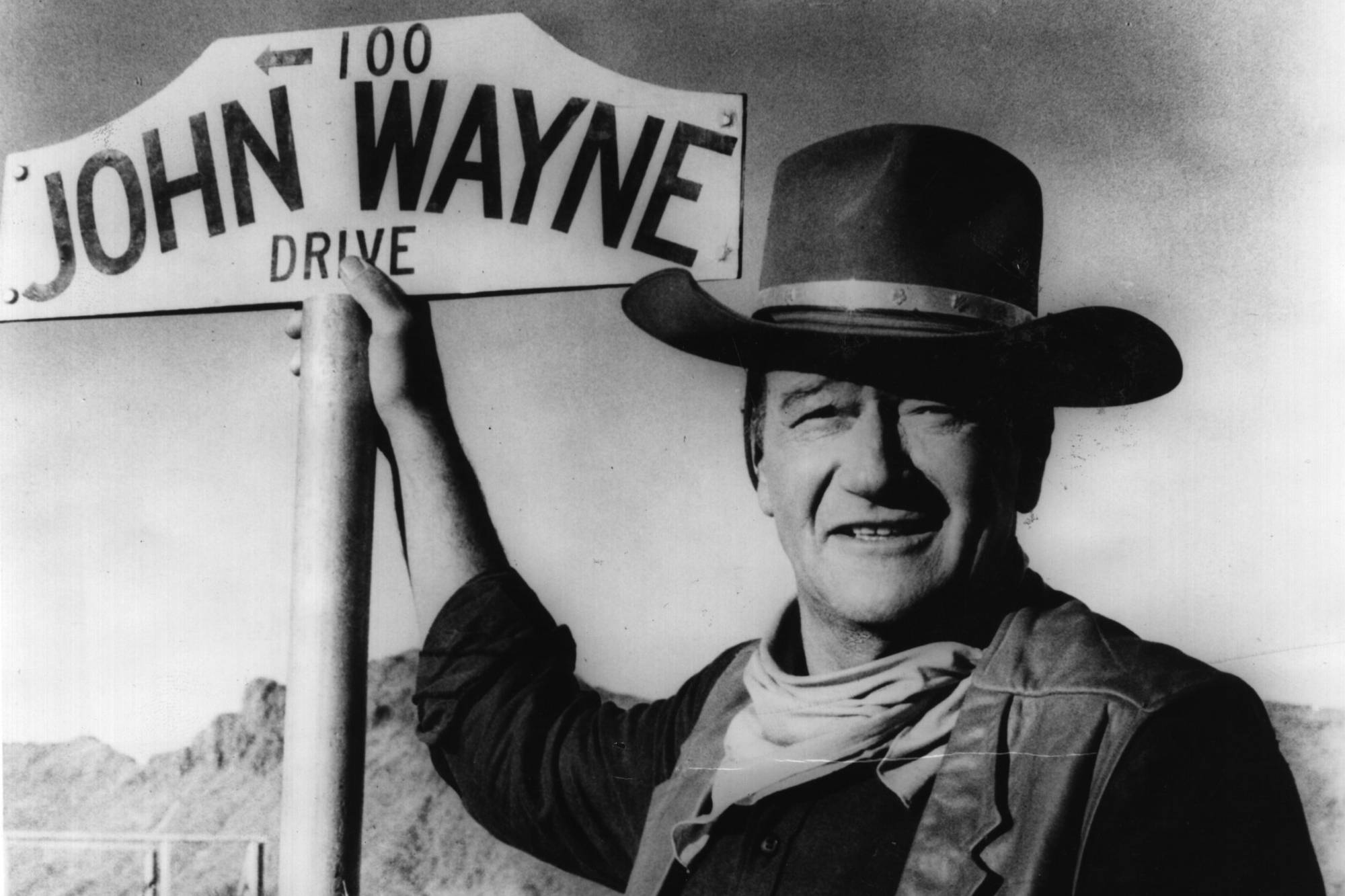 Western movie star John Wayne wearing a Western costume, with his hand on a street sign that reads 'John Wayne Drive'