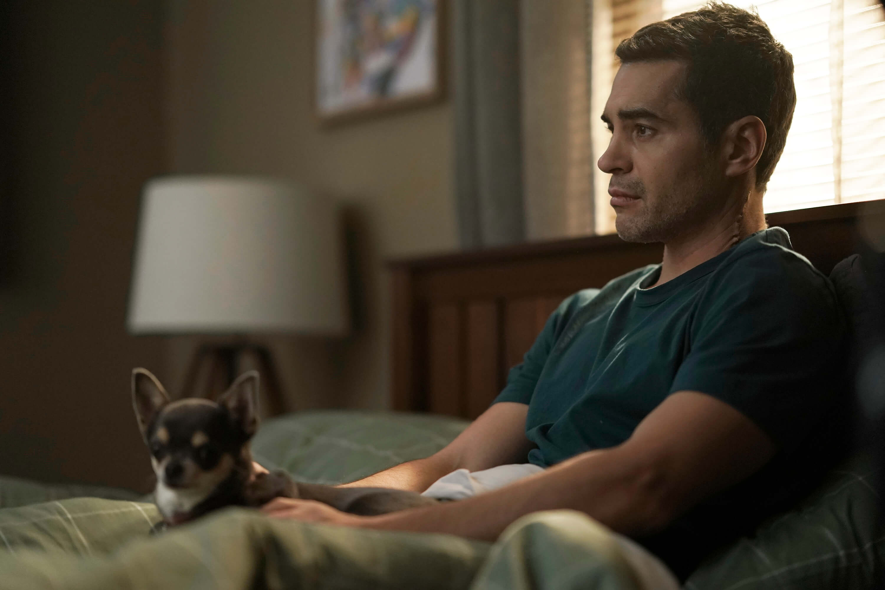 Ramón Rodríguez, in character as Will Trent in the new series 'Will Trent' on ABC, wears a blue shirt while sitting in bed with his chihuahua Betty on his lap.