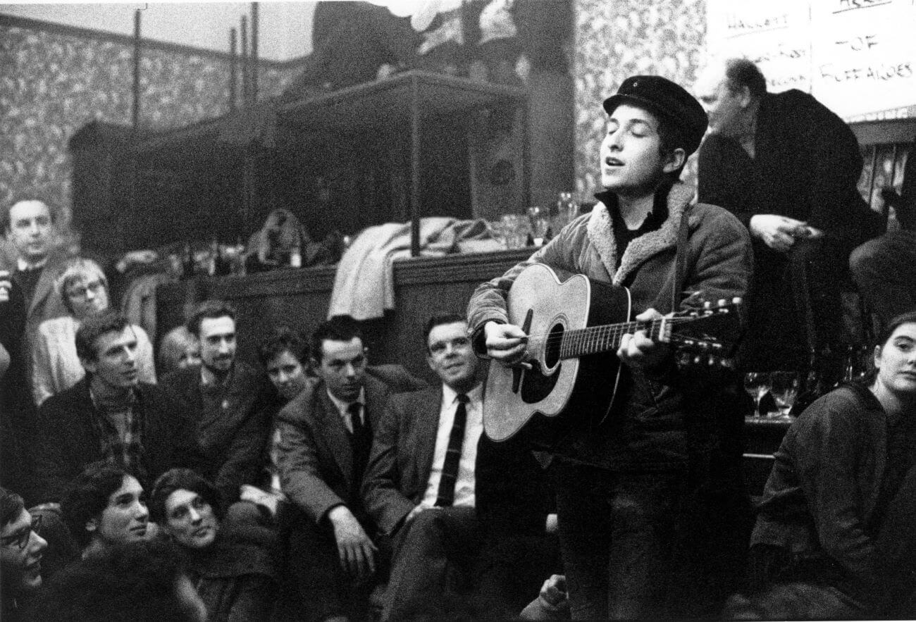 A black and white picture of Bob Dylan wearing a hat and playing guitar to a crowd.