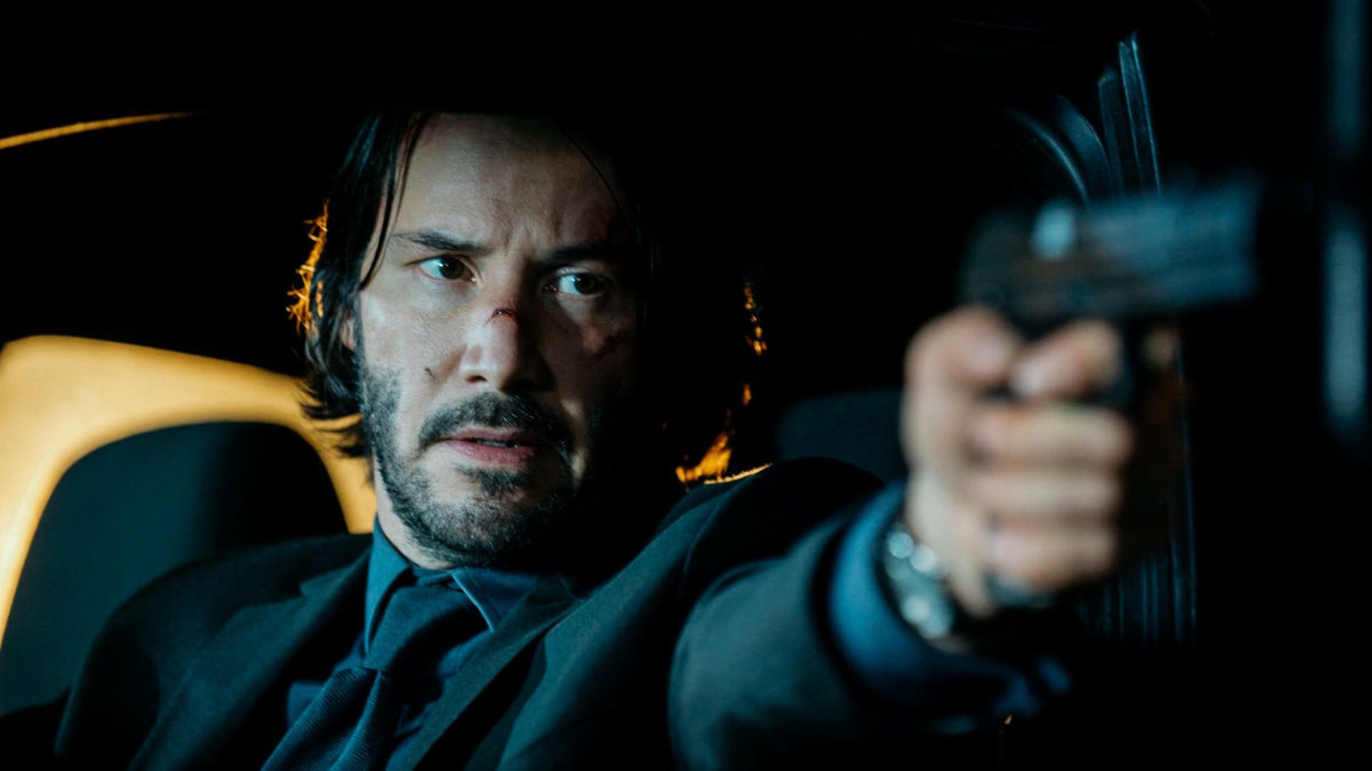 'John Wick' movies ranked Keanu Reeves as John Wick. He's sitting in a car, holding a pistol outward with cuts on his face.