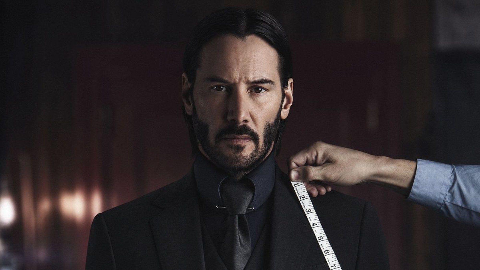 'John Wick: Chapter 2' Keanu Reeves as John Wick looking straight ahead wearing a suit with a hand in the frame tailoring the suit with a measuring tape.
