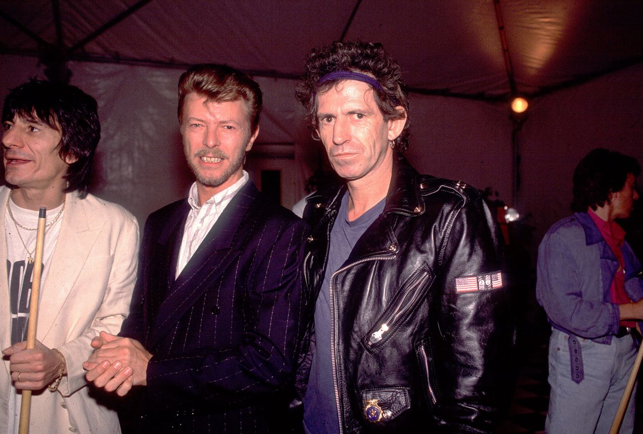 Ronnie Wood holds a pool cue while standing next to Davie Bowie and Keith Richards.