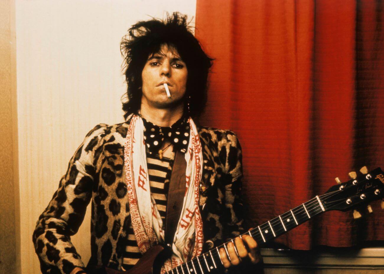 Keith Richards holds a cigarette in his mouth and wears a leopard print shirt. He holds a guitar.