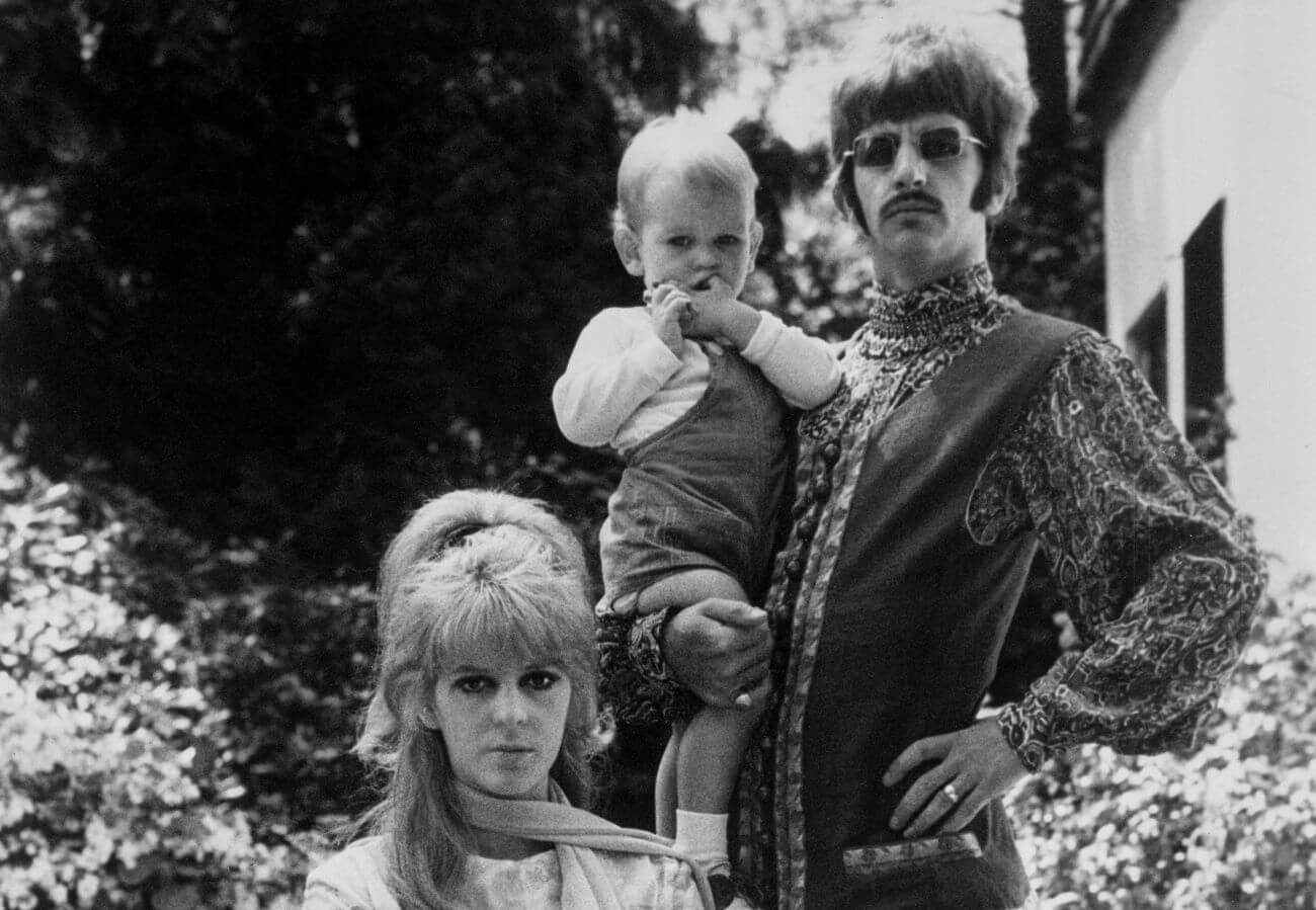 A black and white photo of Maureen Starkey sitting, and Ringo Starr standing and holding his son Zak Starkey.