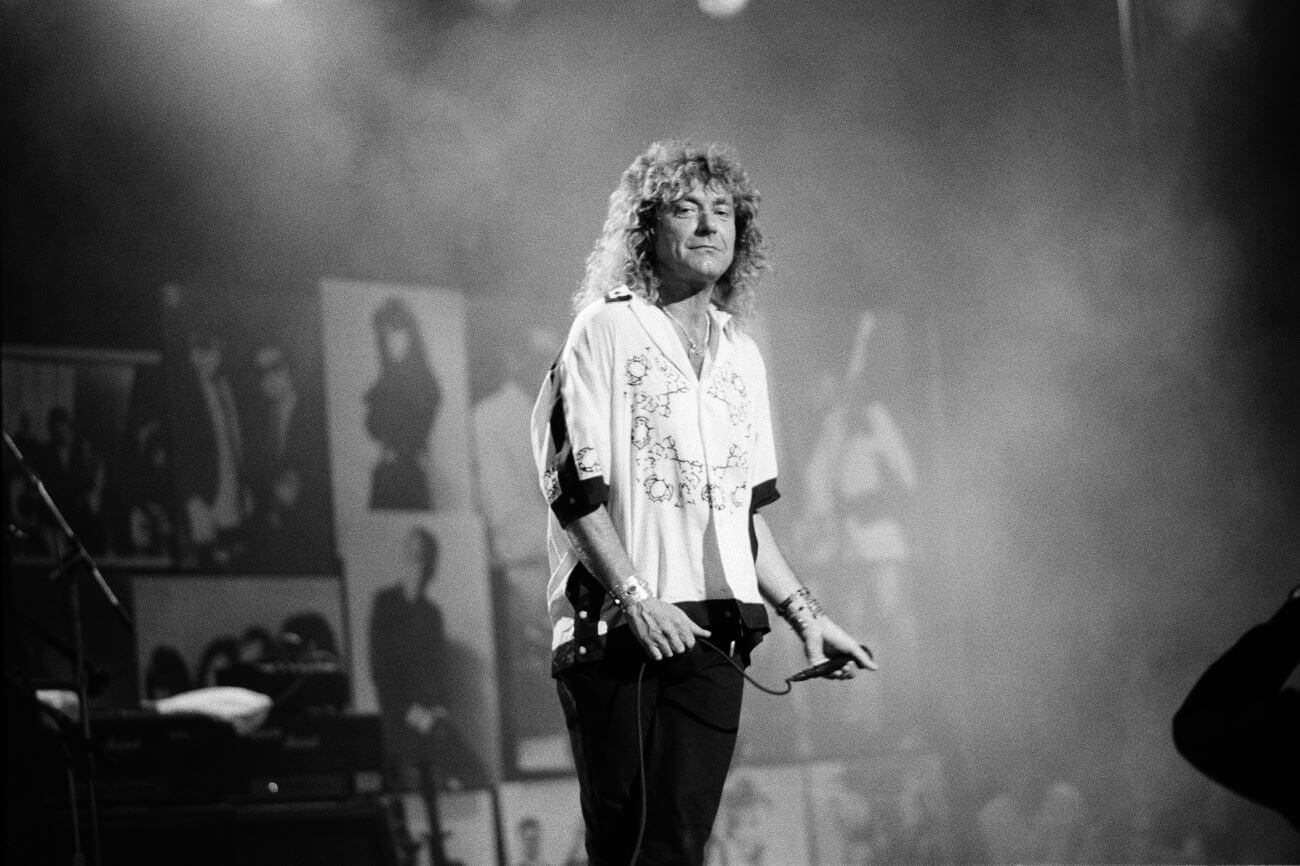 A black and white picture of Robert Plant holding a microphone and standing on a stage.